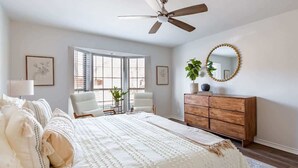 Un dormitorio con una cama, ventilador de techo, cómoda, una silla y una ventana.
