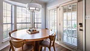 Un comedor con una mesa redonda, sillas, una lámpara colgante y ventanas grandes con persianas.