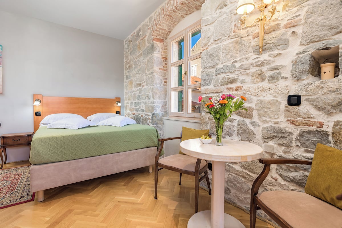 Un dormitorio con pared de piedra, cama de madera, mesita de noche, una mesa redonda con sillas y un florero con flores.