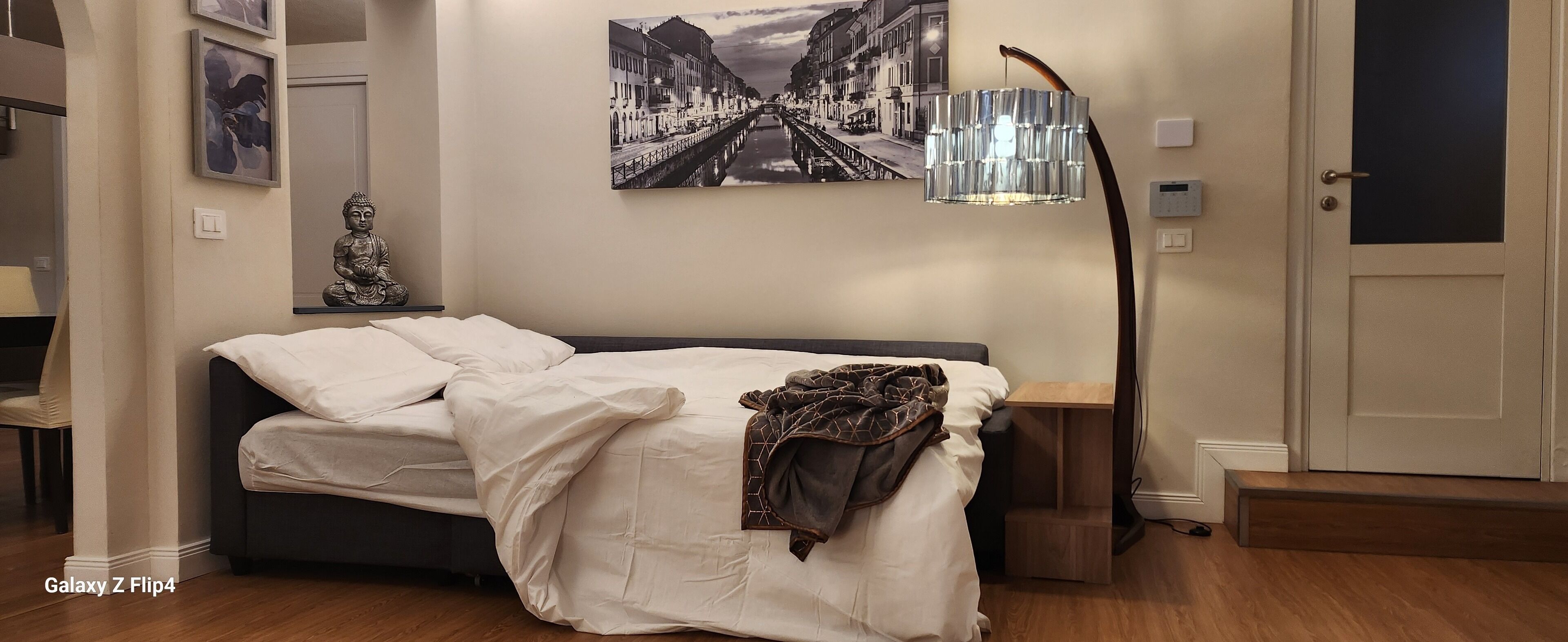 Een slaapkamer met een Galaxy Z Flip4 bed, een Boeddhabeeld, een ingelijst stadsgezicht en een moderne vloerlamp.