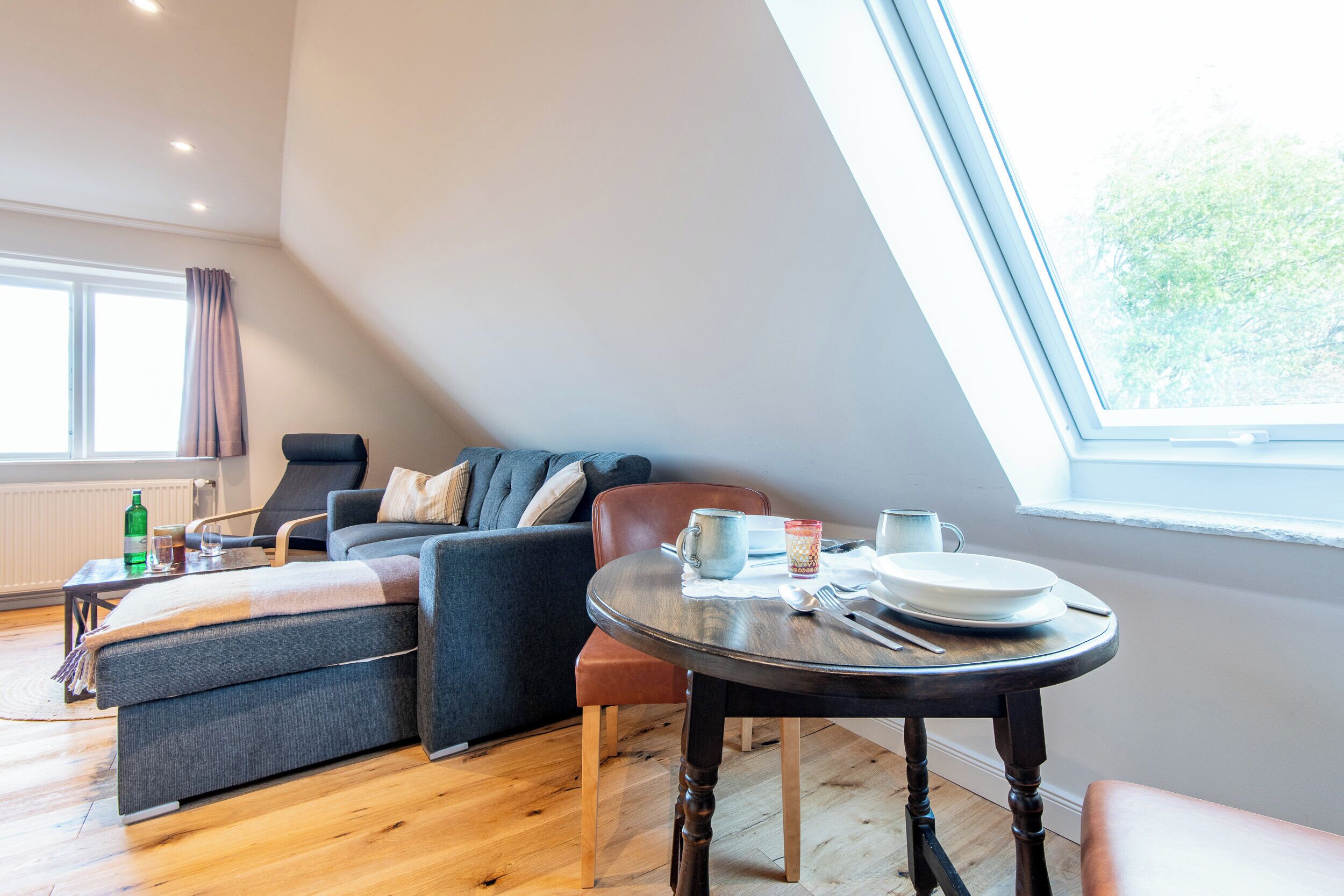 Un salon moderne avec un canapé, une table à manger avec de l’assiette et des couverts, et une vue sur la verdure grâce à une grande fenêtre.