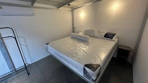 En säng med en madrasskydd, kuddar och ett täcke i ett rum med ett betonggolv och vita väggar.