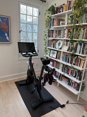 A room with a stationary bike, a bookshelf filled with books, and a window with a view of trees. - Family-friendly Philly Rowhome with Garage and Deck - Dream Location!! (Philadelphia)