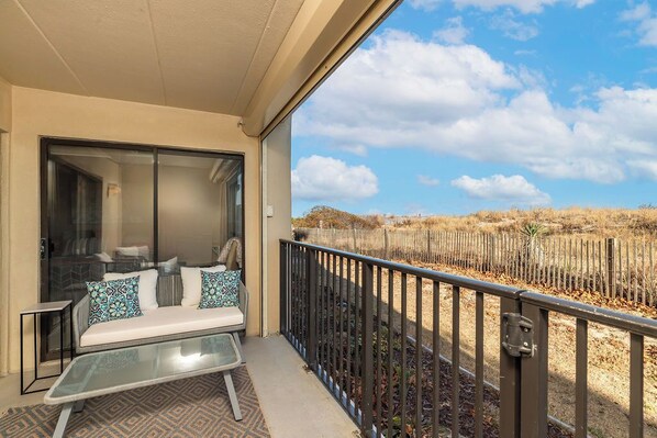 A balcony with a glass table and a sofa, a view of a fence and trees, and a clear sky. - Cozy, comfortable 2 bedroom oceanfront condo with coastal decor, free WiFi, and view of the ocean and dunes just steps to the beach! (Ocean City)