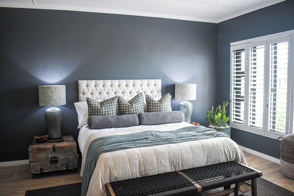 A bedroom with a tufted headboard, a large bed with multiple pillows, bedside lamps, a rustic chest of drawers, a bench, and a window with shutters.