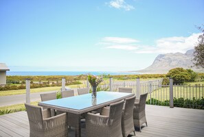A deck with wicker furniture and a glass table, overlooking a coastal landscape with a mountain in the distance.