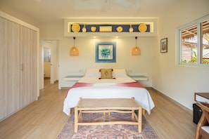 A bedroom with a bed, a bench, a patterned rug, a shelf with decorative items, and a window with a view of the outdoors.