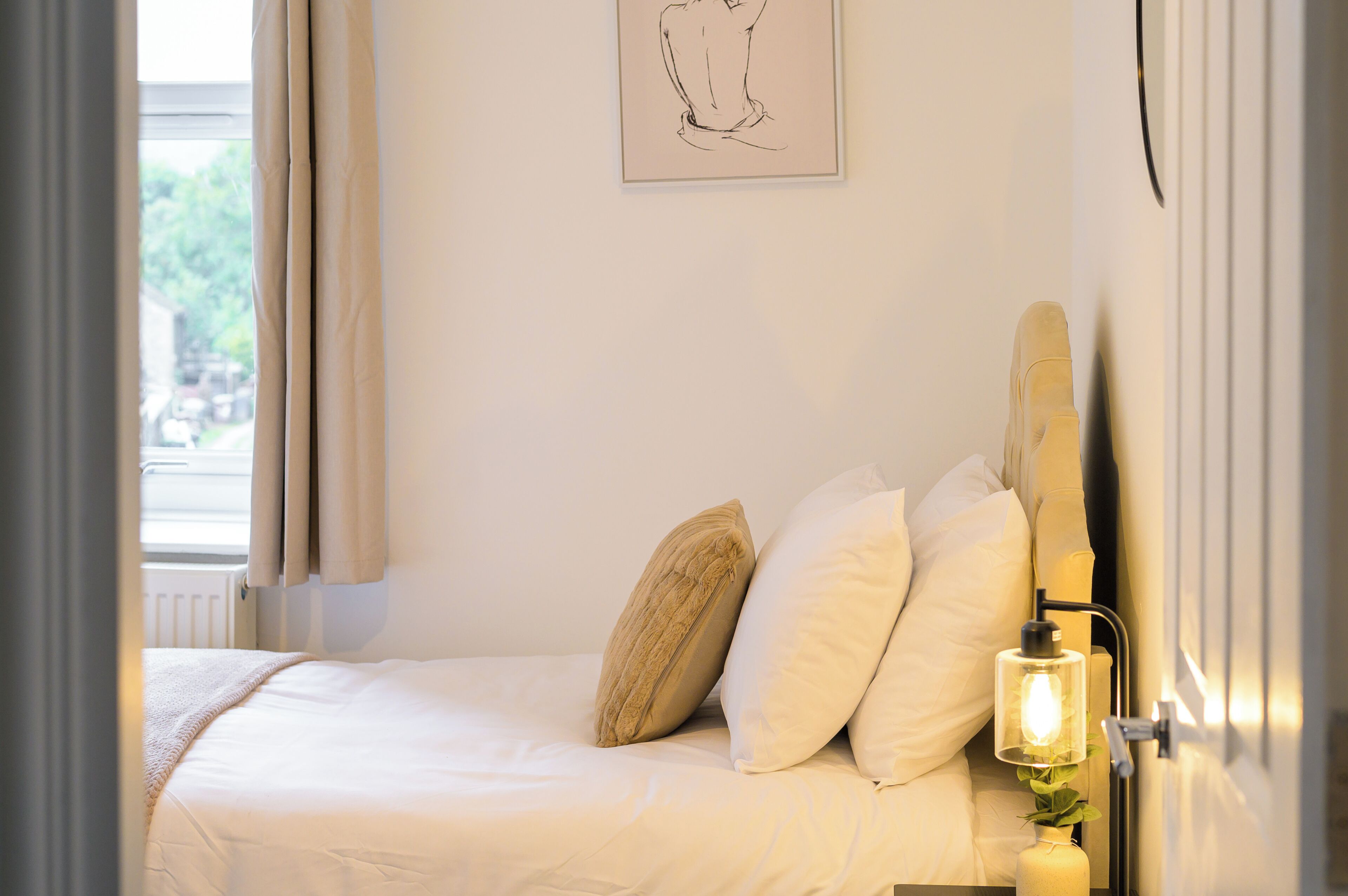 Ein ordentlich bezogenes Bett mit weißen Laken und beigen Kissen, ein Nachttisch mit Lampe und ein gerahmtes Kunstwerk an der Wand.