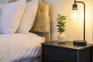 Een net opgemaakt bed met witte lakens, een nachtkastje met een vaas en een lamp, en een donker bijzetmeubel.