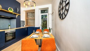 A compact kitchen with a built-in oven, a dining table with orange chairs, and a large wall clock.