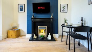 A living room with a fireplace, a TV mounted above it displaying 'NETFLIX', and a dining area with a table and chairs.