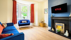 A living room with a blue sofa, a fireplace, a television mounted on the wall, and a window with curtains.