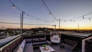 A rooftop terrace with a sofa, chair, and table, overlooking a cityscape at sunset.
