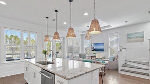 A modern kitchen with a central island, pendant lights, and a large window overlooking a deck. - Pearl House: Luxury Retreat with Bikes and Golf Cart, and Pond & Nature Views, Pickleball Courts! (Santa Rosa Beach)