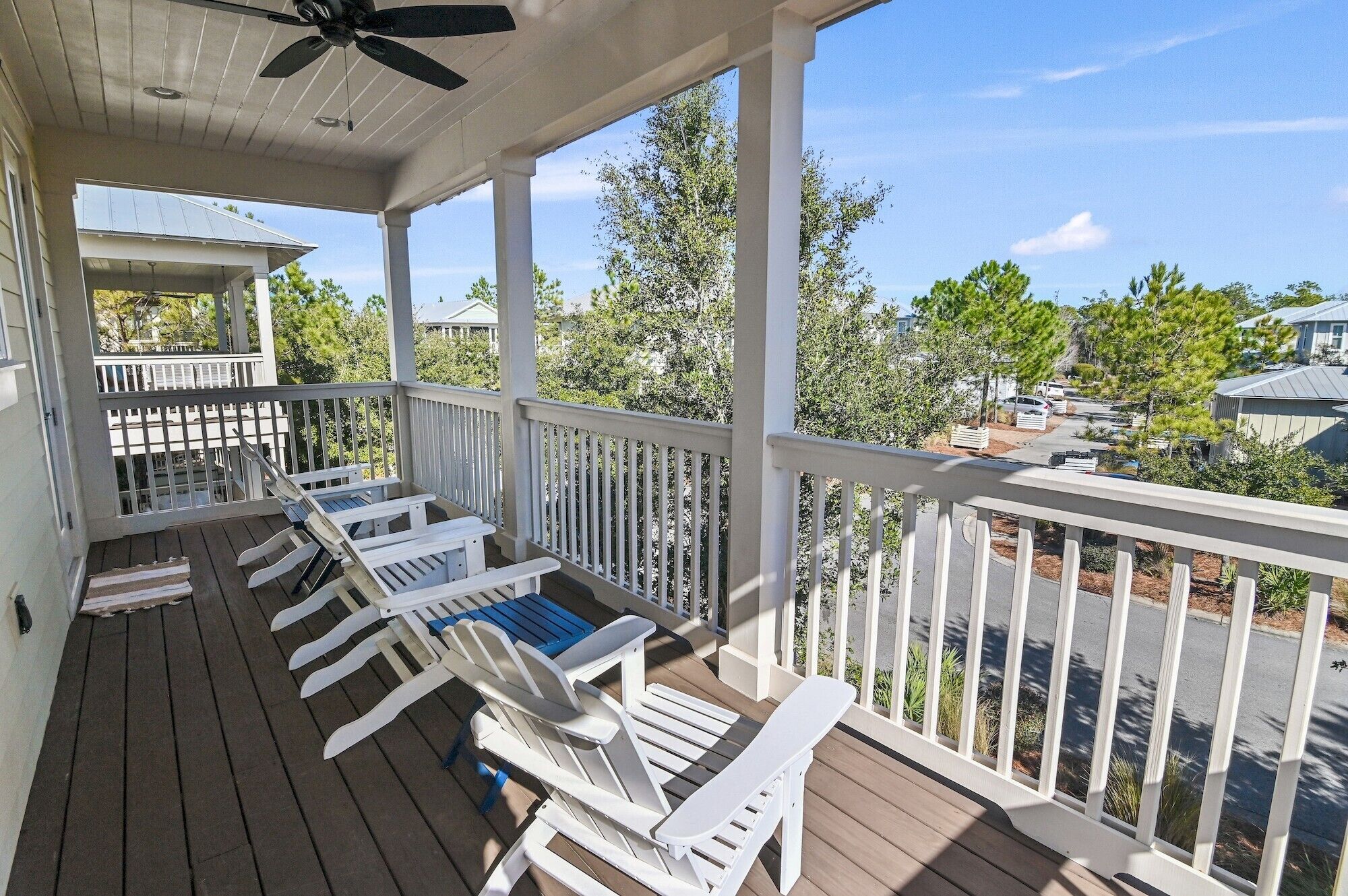 A covered porch with white railings and wooden flooring, featuring several white Adirondack chairs and a ceiling fan.