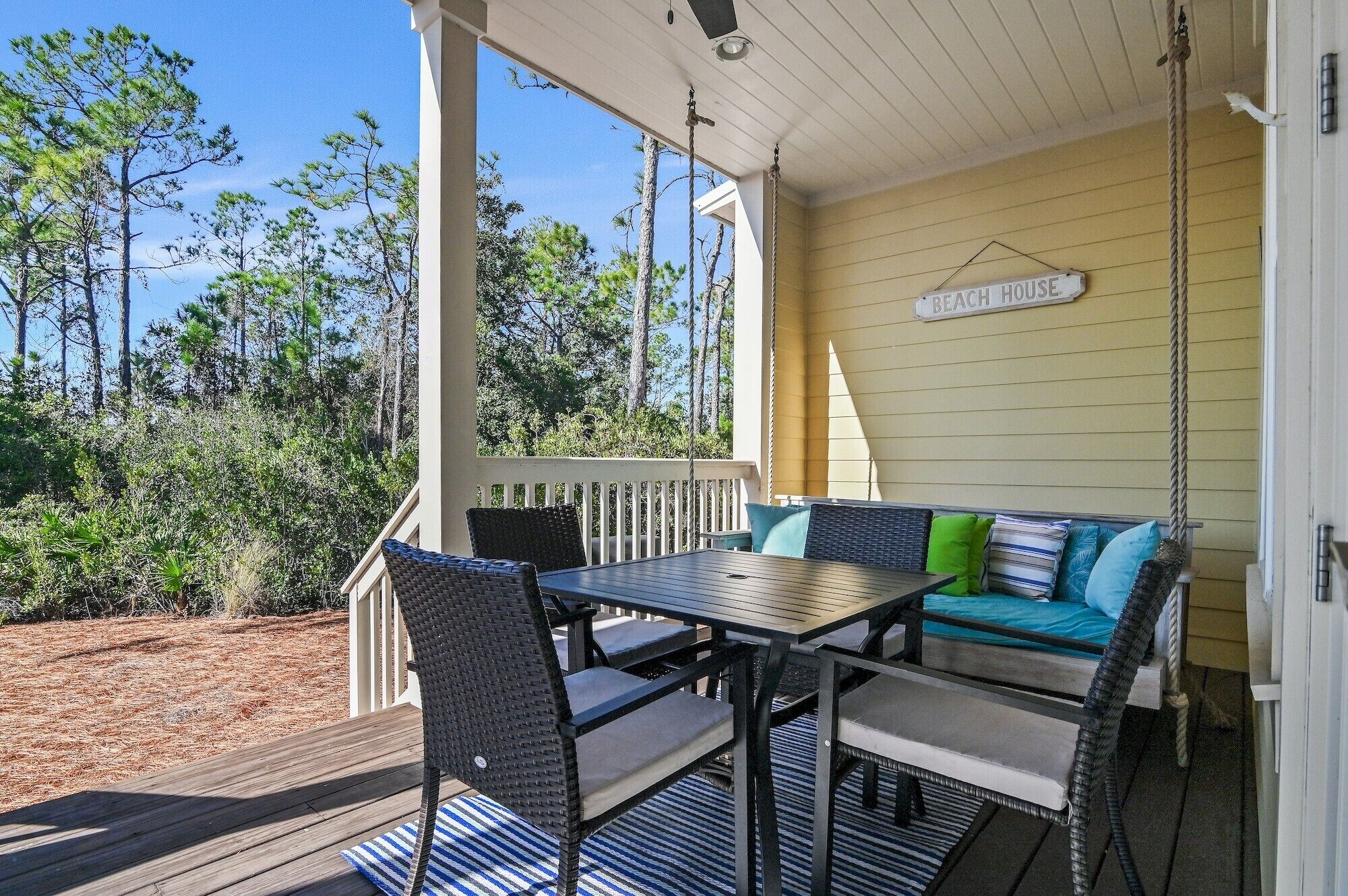 A covered porch with a table and chairs, a sofa with cushions, and a sign reading "BEACH HOUSE".