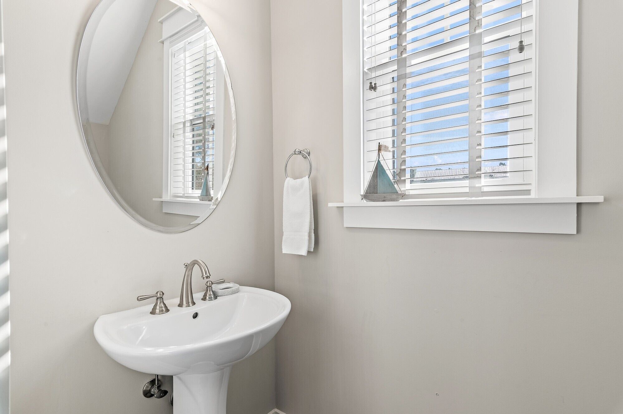 Une salle de bain avec un lavabo blanc, un miroir rond et une fenêtre avec des stores.