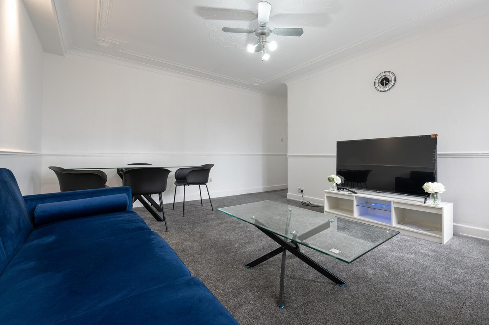 Un salon moderne avec un canapé bleu, une table basse en verre, un téléviseur à écran plat posé sur un meuble blanc et un ventilateur de plafond.