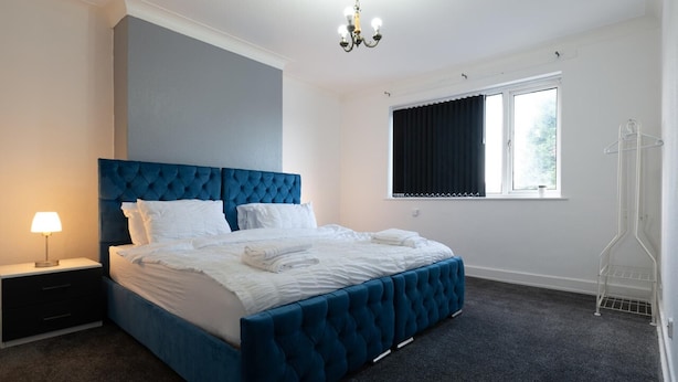 A bedroom with a large bed, a nightstand with a lamp, a window with curtains, and a radiator. - 2-bedroom- Stoke-on-trent-sleeps 5-petsok (Stoke-on-Trent)