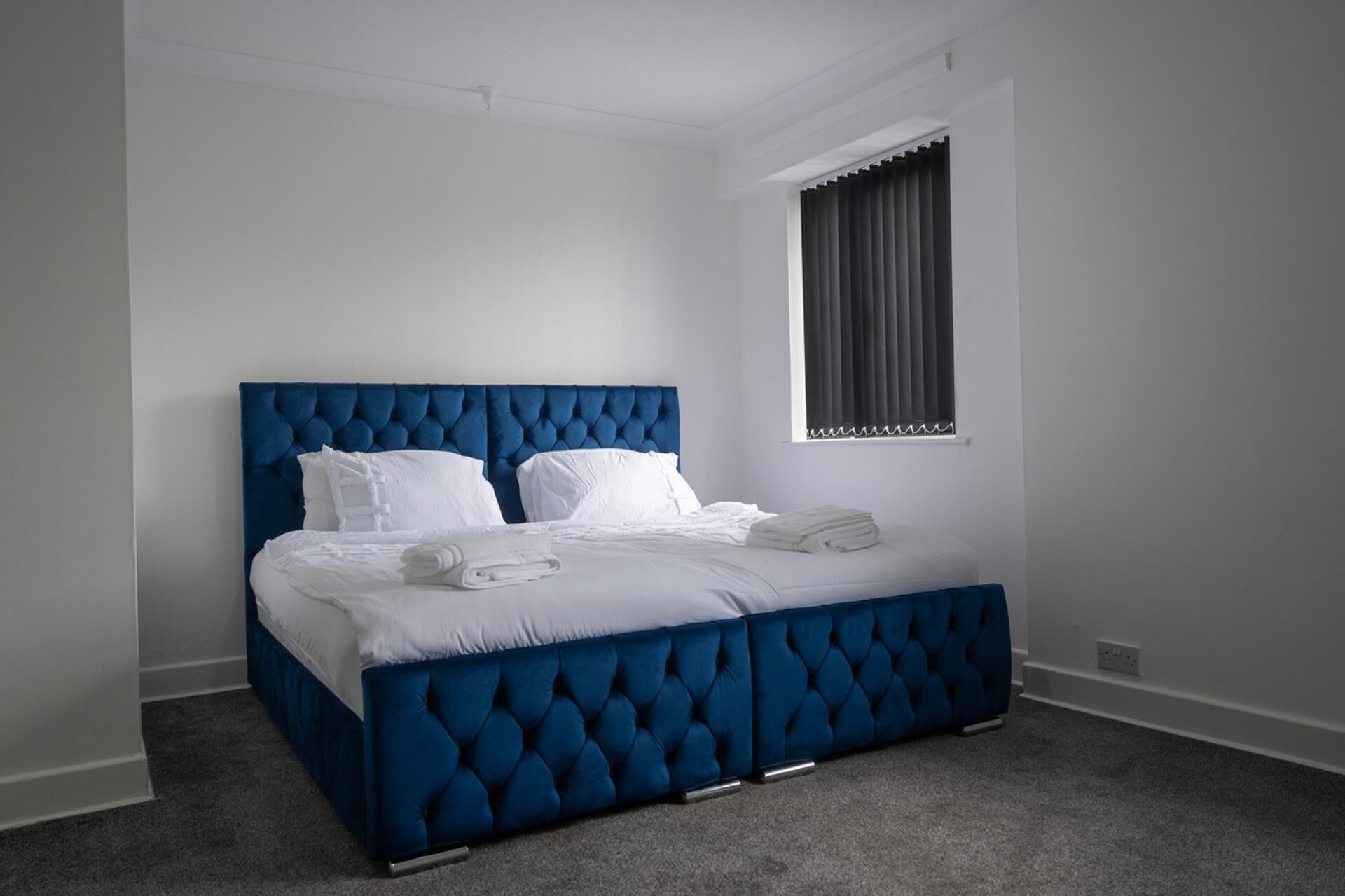 A bedroom with a blue tufted headboard, white bedding, and a window with dark curtains.
