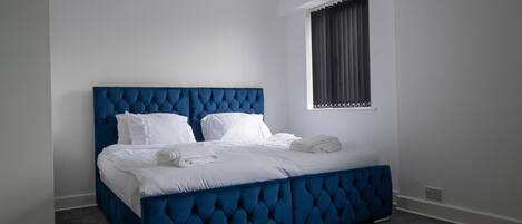 A bedroom with a blue tufted headboard, white bedding, and a window with dark curtains.