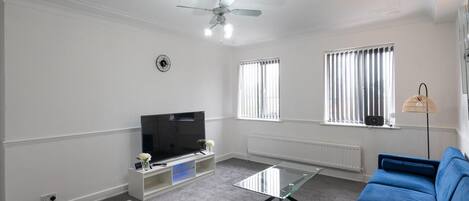 Una sala de estar con un televisor de pantalla plana, un ventilador de techo, un sofá azul y una mesa de centro de vidrio.