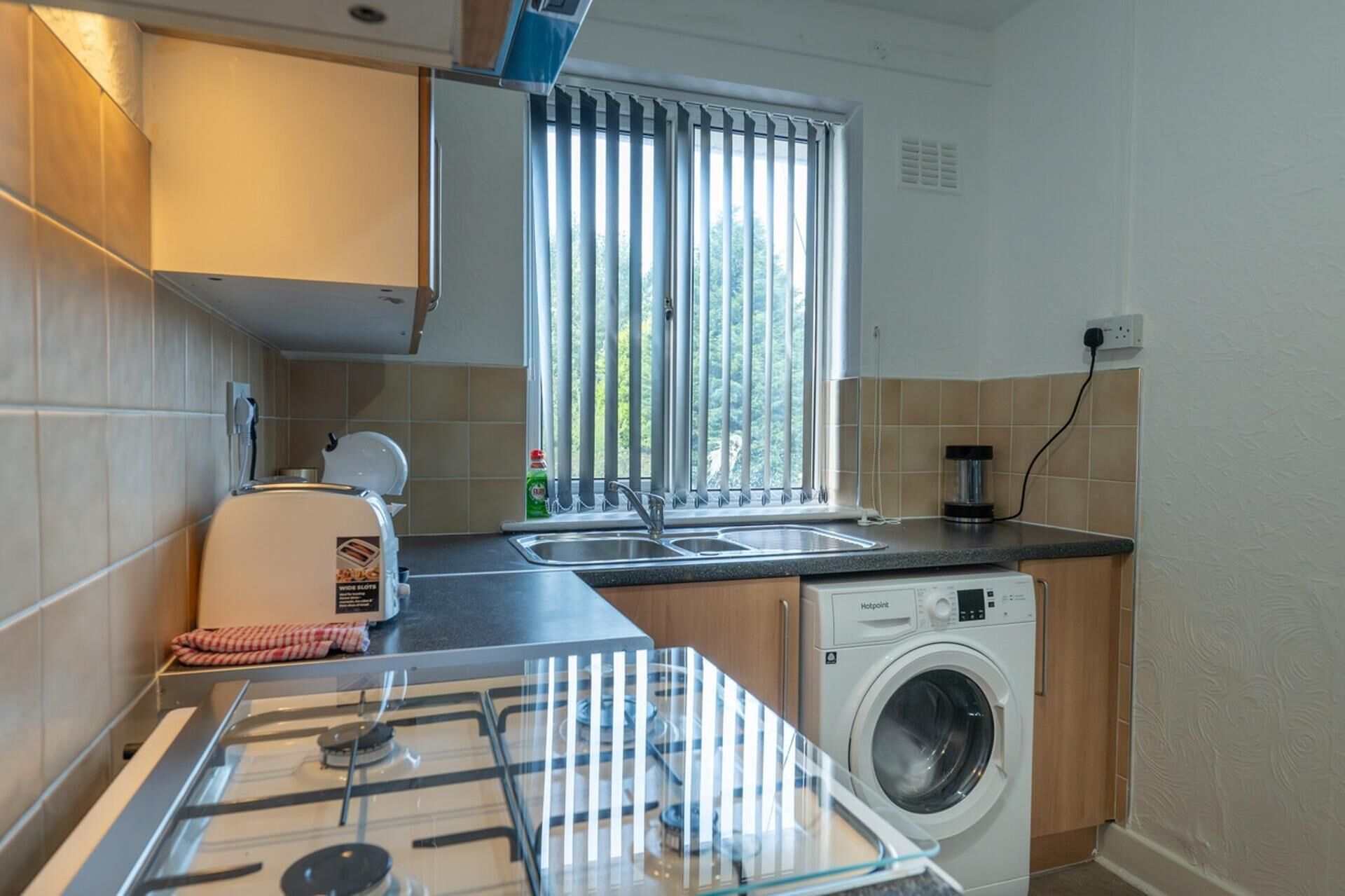 Une cuisine compacte équipée d’une cuisinière à gaz, d’un lave-linge, d’un évier et d’une fenêtre avec des stores.