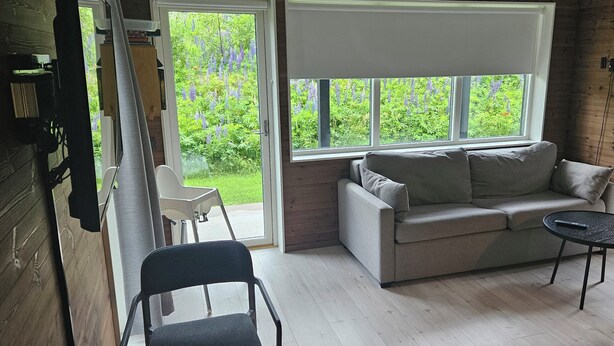 A cozy room with a wooden floor, a grey sofa, a small round table, and a window with a view of greenery. - Bergsgården Lodge Järvsö (Jarvso)
