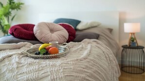 Une chambre d’hôtel avec un lit sur lequel est posé un plateau décoratif rempli de fruits, une lampe de chevet et une plante.