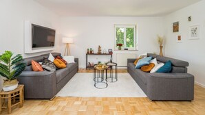 Un salon moderne avec deux canapés gris, un tapis blanc, un plancher en bois, un téléviseur fixé au mur, une petite table agrémentée d’objets décoratifs et une fenêtre donnant sur des arbres.