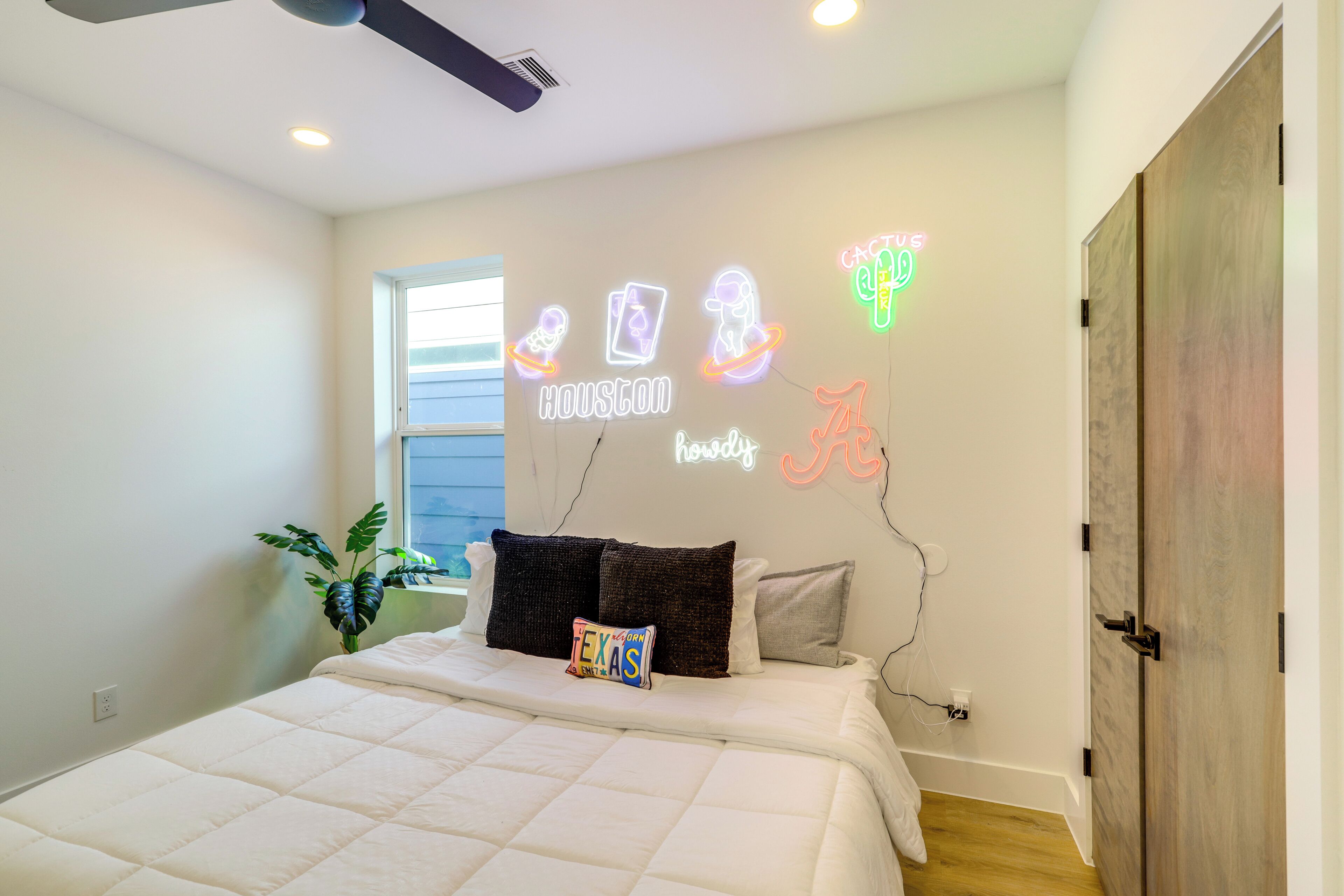 Ein Schlafzimmer mit Bett, Kissen, einer Neonleuchtreklame, einem Fenster und einer Pflanze.