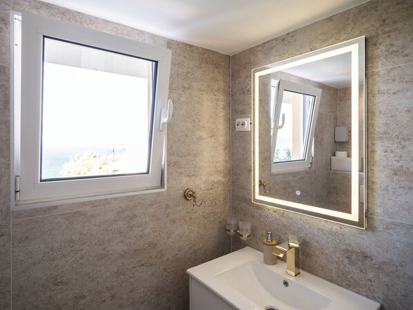 A bathroom with a large window, a sink with a gold faucet, and a mirror with a light.