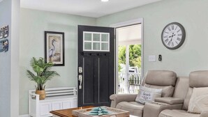A living room with a dark door, a beige sofa, a wooden coffee table, and a wall clock. - Seashell Retreat 3/2 By Beach Star (Murrells Inlet)