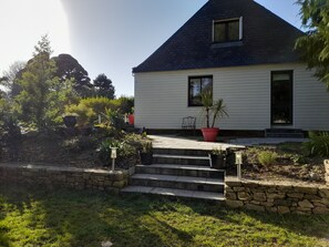 Situated on a sloped terrain, this two-story house features a steeply pitched roof and a garden with diverse plant species.
