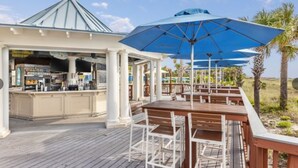 Un'area esterna coperta con un bar, tavoli di legno e ombrelloni blu.