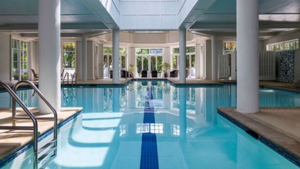 Una piscina interna con acqua cristallina e blu, circondata da colonne bianche e ampie finestre.