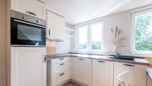 A modern kitchen with white cabinetry, a built-in oven, a window with a view of greenery, and a white chair. - Vogelkoje Living in nature, vacation apartment with terrace and beach chair (Rodenäs)