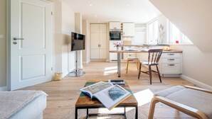 A modern kitchen with white cabinets, a dining area with a wooden table and chairs, and a TV on a stand. - Vogelkoje Living in nature, vacation apartment with terrace and beach chair (Rodenäs)