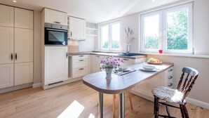 A modern kitchen with white cabinetry, a built-in oven, a wooden island with a vase of flowers, and a window providing natural light. - Vogelkoje Living in nature, vacation apartment with terrace and beach chair (Rodenäs)