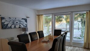 Un coin repas avec une table en bois, des chaises noires, un tableau au mur et de grandes fenêtres avec des rideaux.