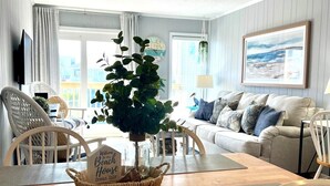 Una sala de estar con una mesa de comedor de madera, sillas blancas, un sofá gris y un ventanal con vistas a edificios.