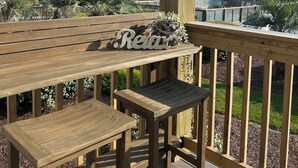 Una terraza de madera con un banco y una mesa, un letrero que dice "Relax" y vista al océano con una casa al fondo.