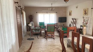 Uma sala de estar ampla com piso de azulejos, uma área de refeições com mobiliário de madeira e uma grande janela com cortinas.