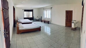 Quarto com duas camas de madeira, uma pequena mesa, uma cadeira, janela com cortinas, porta e aquecedor.
