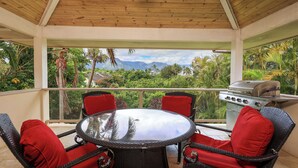 Een overdekt terras met een ronde tafel en vier stoelen, een barbecue en uitzicht op een weelderig landschap met bergen.
