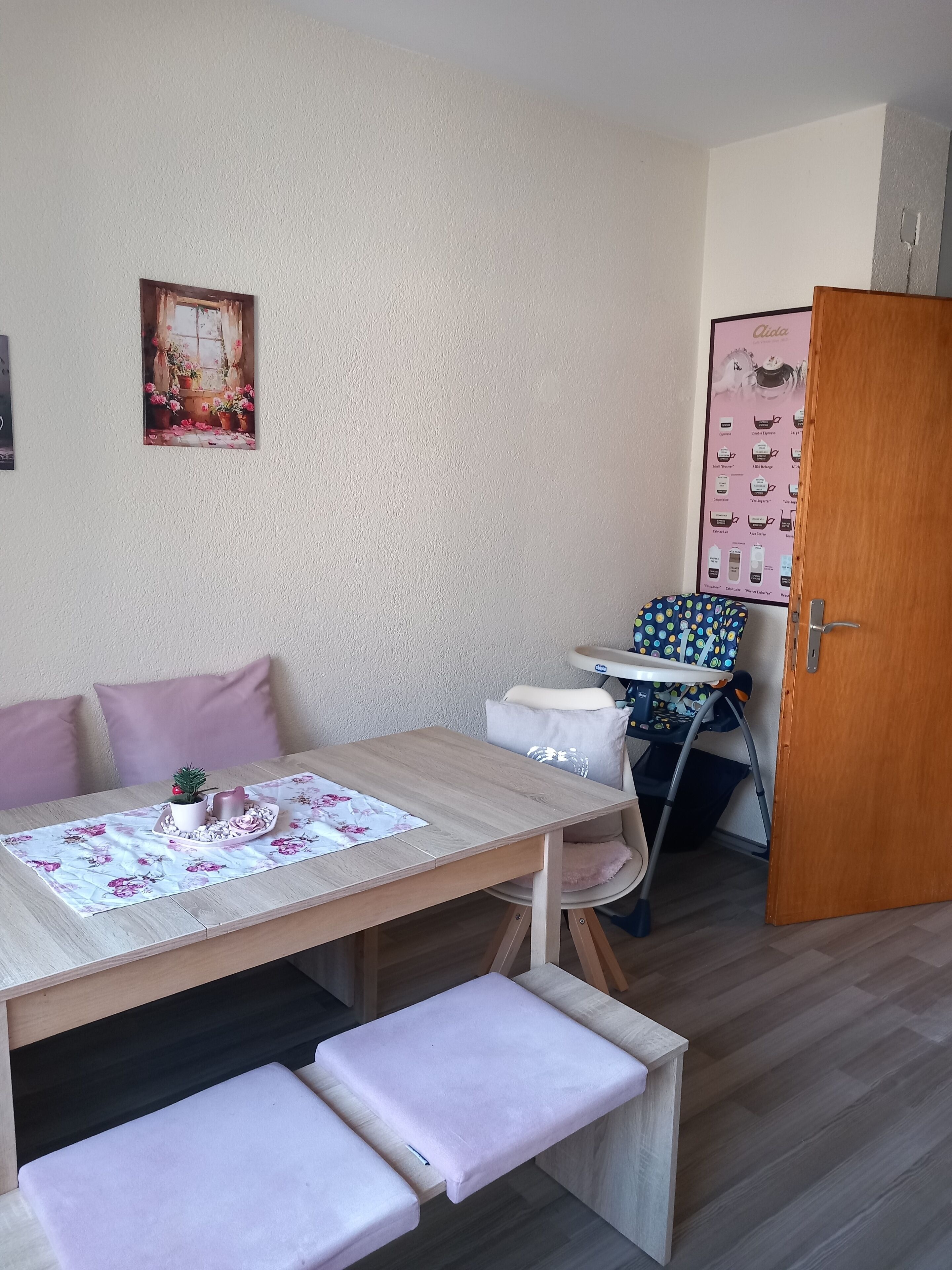 A small room with a wooden table, two benches, a chair, a door, and a poster on the wall.