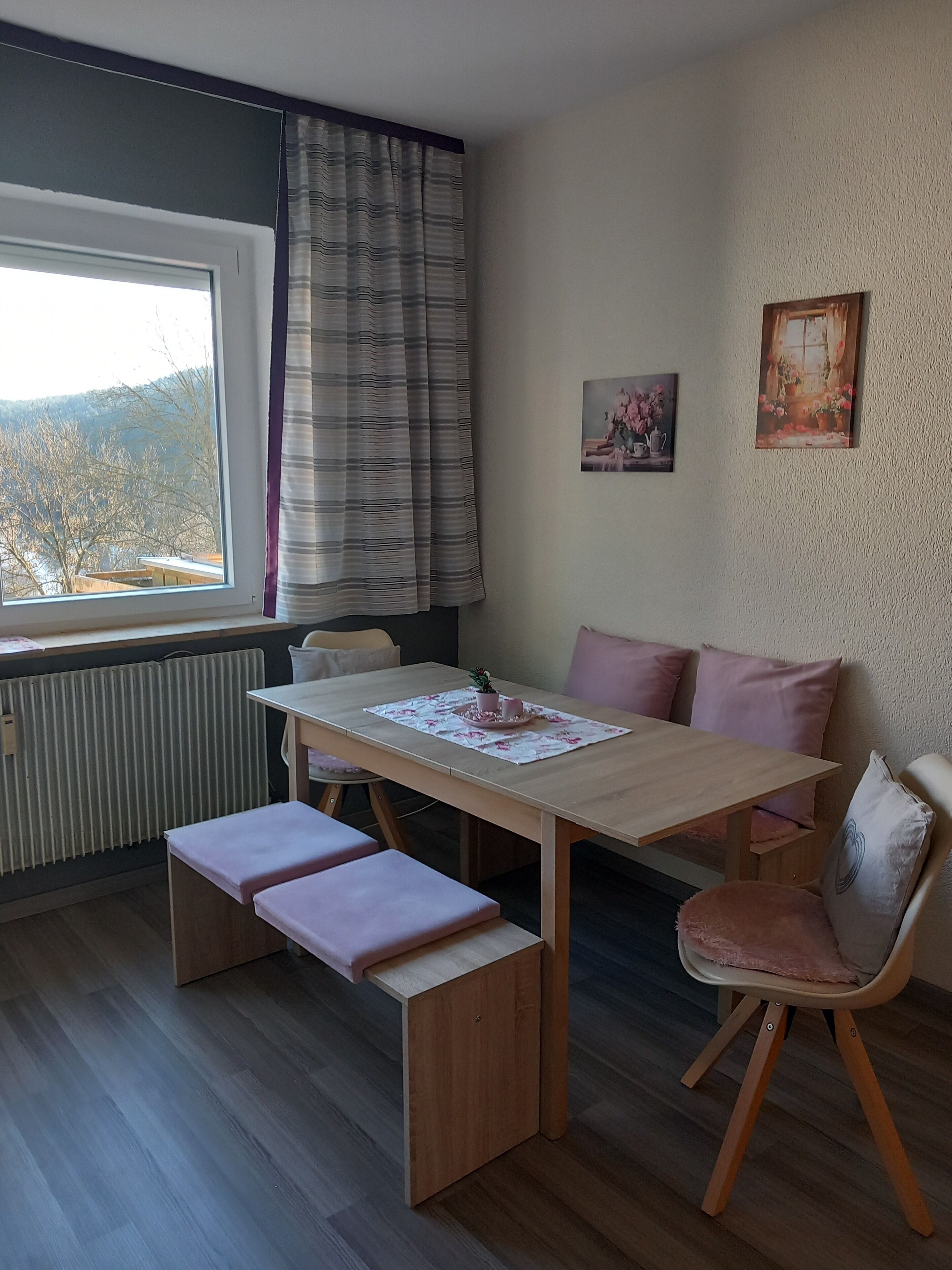 Una habitación con una mesa de madera, un banco y un sofá con cojines. Hay una ventana con cortinas, un radiador y dos cuadros en la pared.