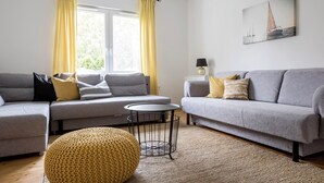 Una sala de estar moderna con dos sofás grises, un taburete amarillo tejido, una mesita redonda y un cuadro de veleros en la pared.