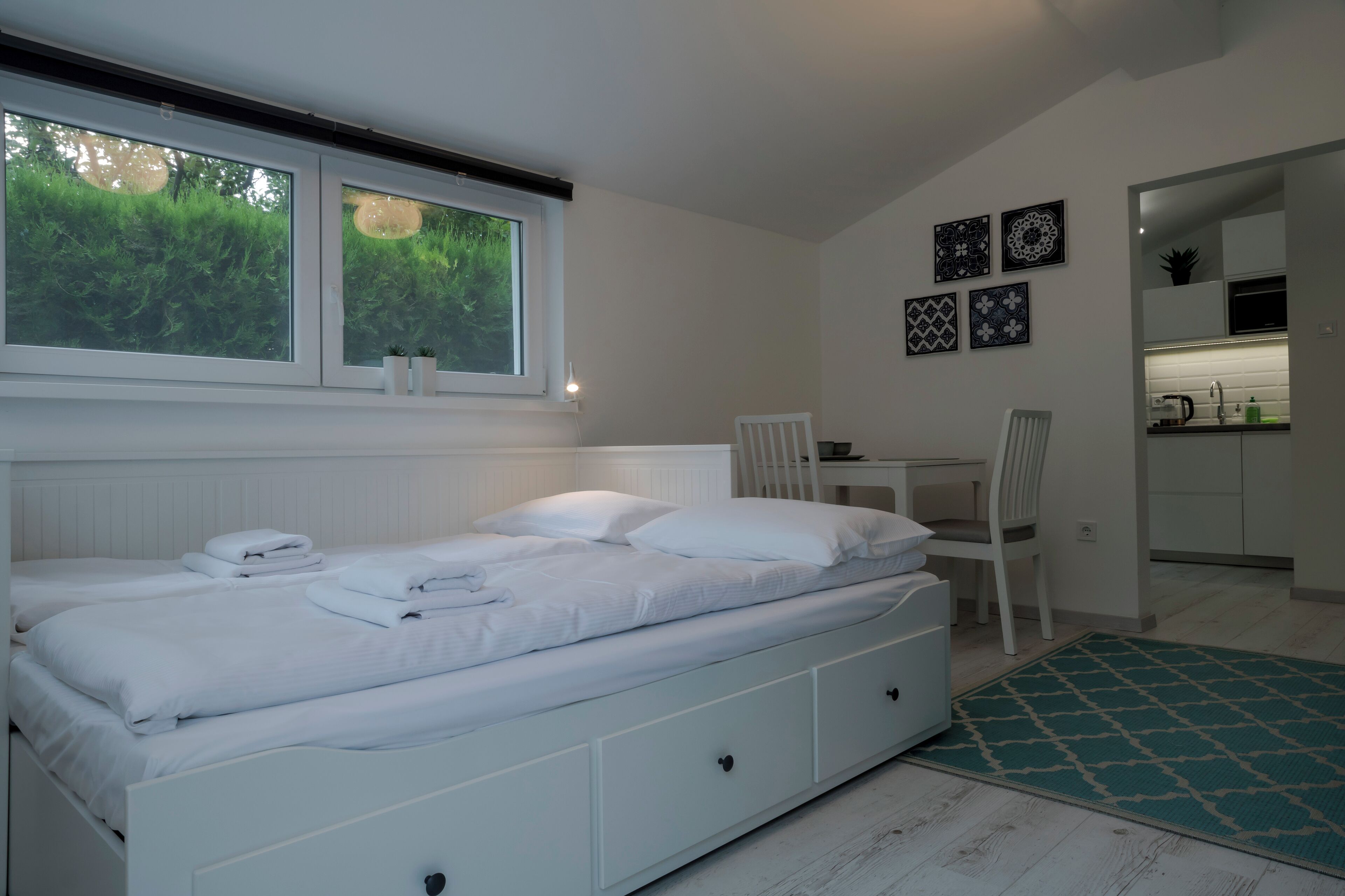 Een slaapkamer met een bed, een bureau en een stoel. Er is een raam met zonwering, een radiator en een kleine plant op de vensterbank.
