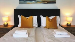 Una cama doble con ropa blanca, dos almohadas amarillas mostaza y un cuadro con barcos a vela encima.